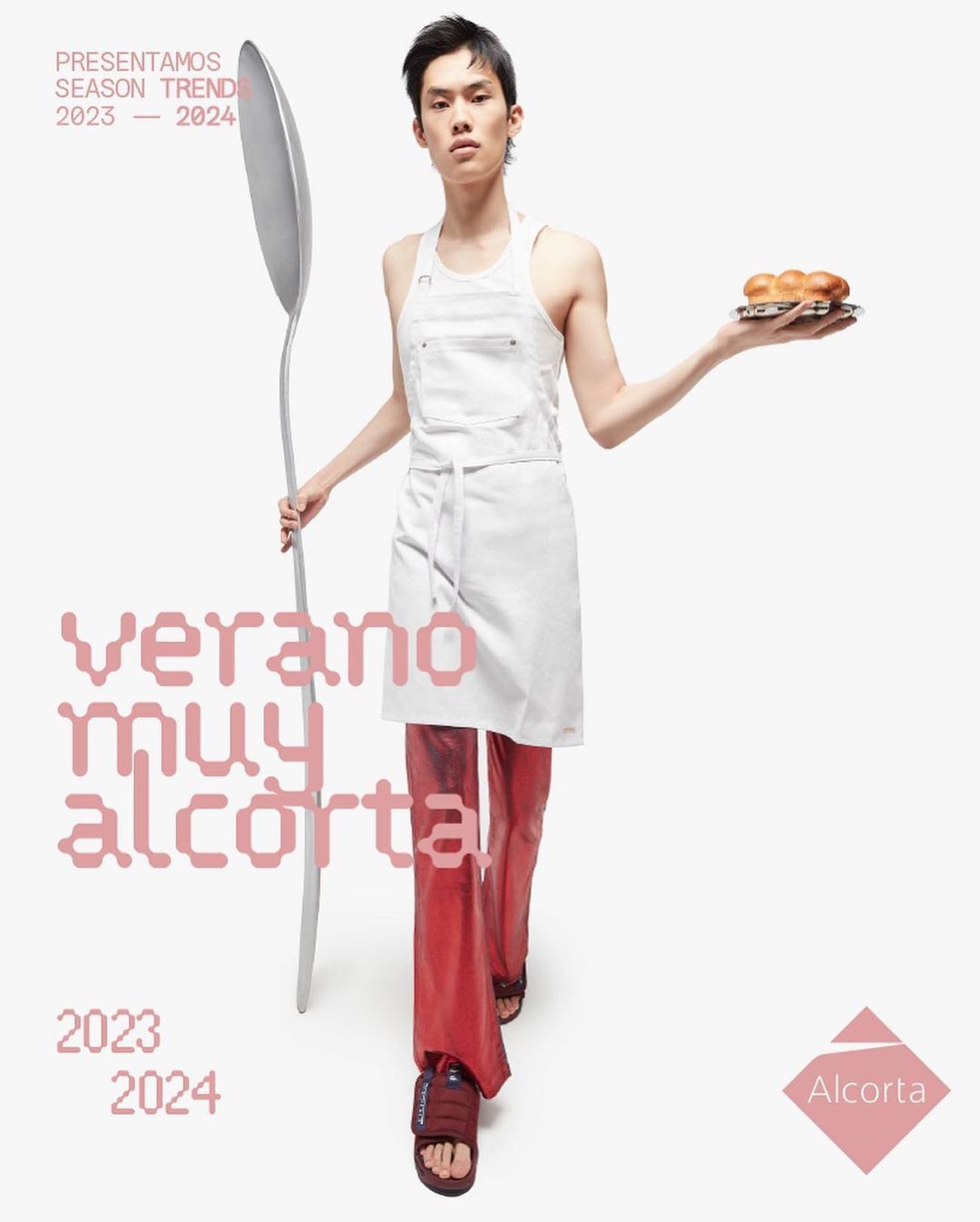 Asian man poses indoors wearing an apron and red pants while holding a plate of bread and a large spoon for a fashion advertisement.