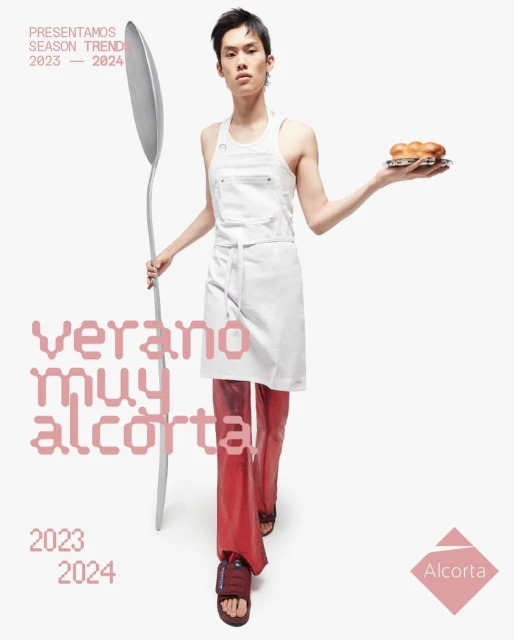 Asian man poses indoors wearing an apron and red pants while holding a plate of bread and a large spoon for a fashion advertisement.