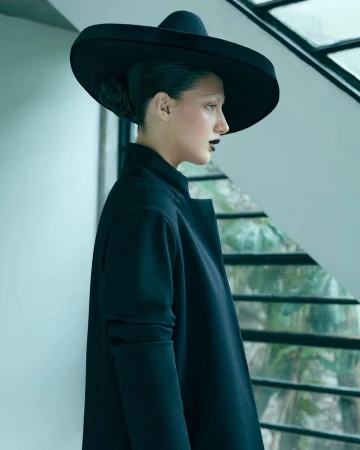 Woman in black hat and coat stands near window. Dark lipstick and swept back hair. Side view, portrait orientation. Fashion shot.