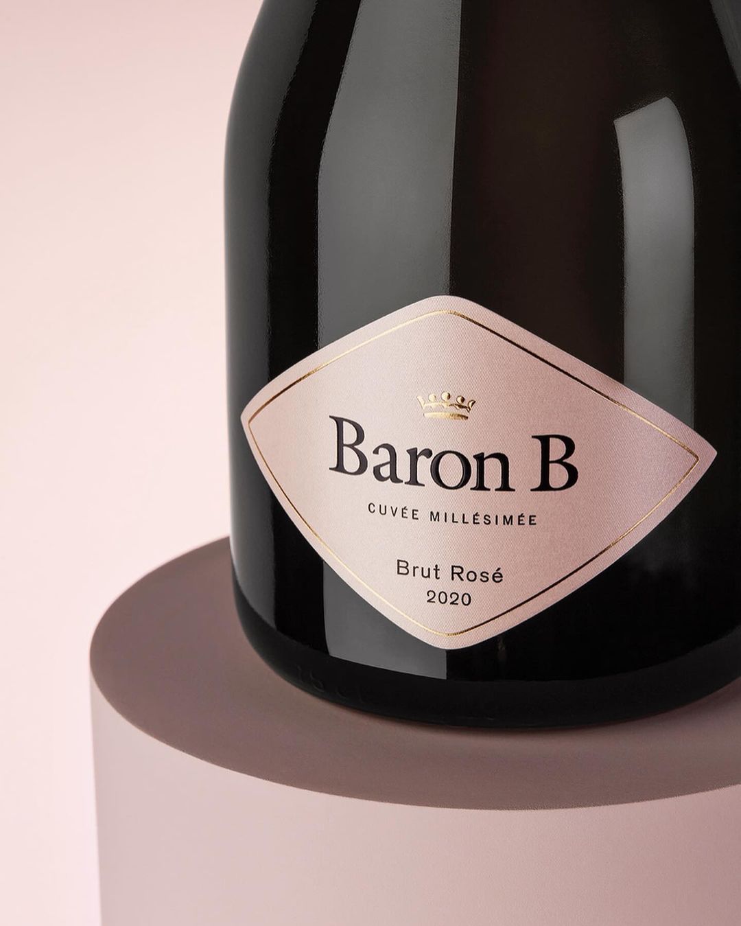 Elegant bottle of Baron B Brut Rosé 2020 champagne with a pink label, displayed on a circular podium against a soft pink background.