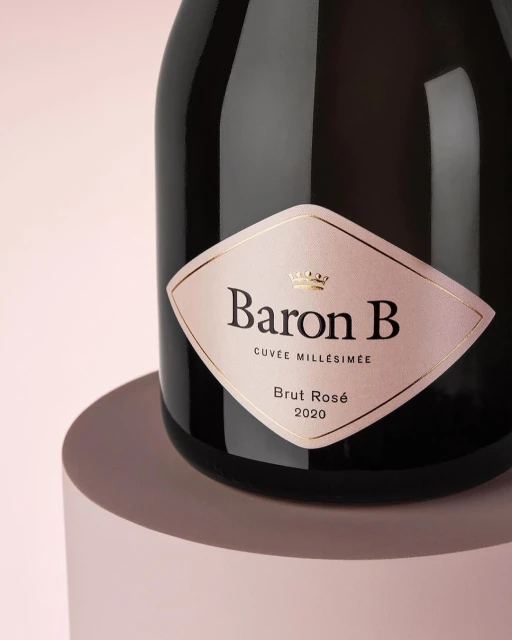 Elegant bottle of Baron B Brut Rosé 2020 champagne with a pink label, displayed on a circular podium against a soft pink background.