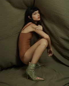 Model with dark hair and bangs crouching in a brown bodysuit and green textured ankle boots against a draped olive backdrop.