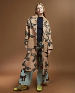 Model in printed bow coat and patterned trousers standing against a warm brown backdrop