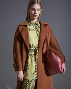 Model wearing a brown coat and patterned green top holding a structured leather shoulder bag against a grey background