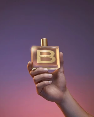 A hand holding a rectangular glass perfume bottle with a golden cap against a gradient purple and pink background