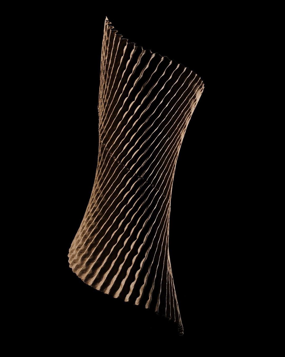 Twisted, vertically-oriented geometric sculpture on black. The sculpture is made of grooved tan material and has a helical shape.