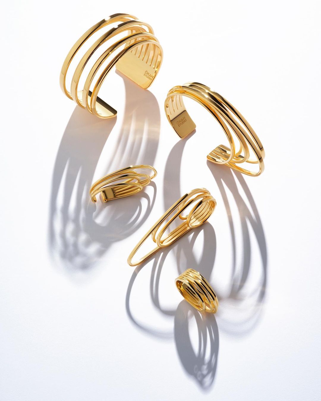 Close-up studio shot of five gold metal cuff bracelets and rings with shadows on a white background. Fashion jewelry product shot.