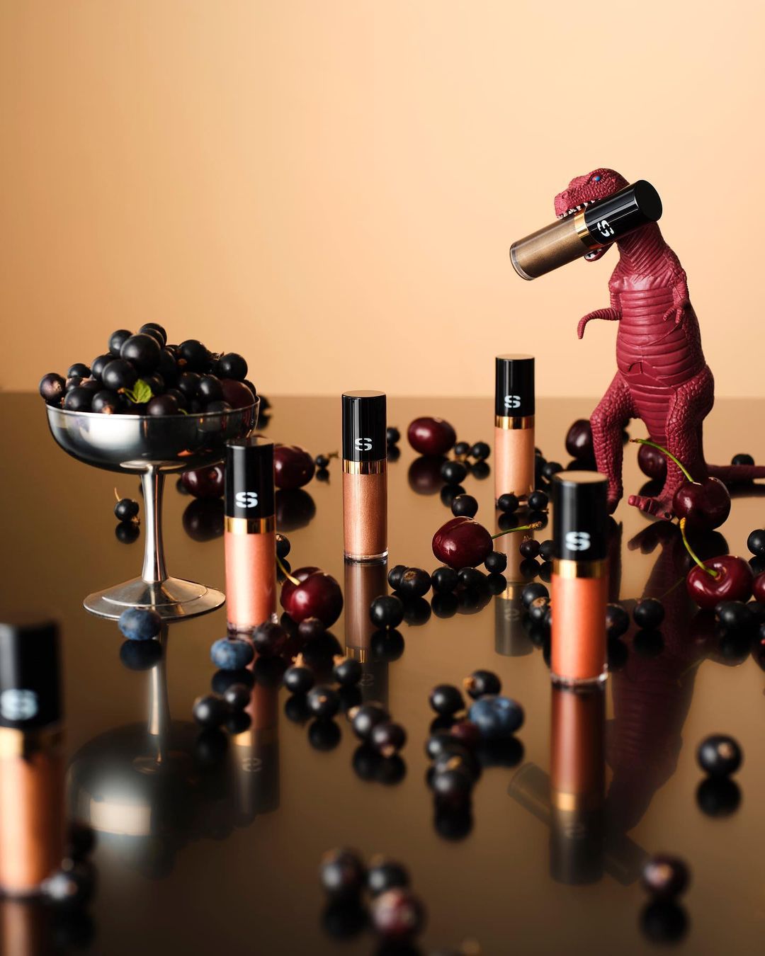 Product shot of cosmetic tubes with berries and a toy dinosaur on a reflective surface in front of a peach-colored background.