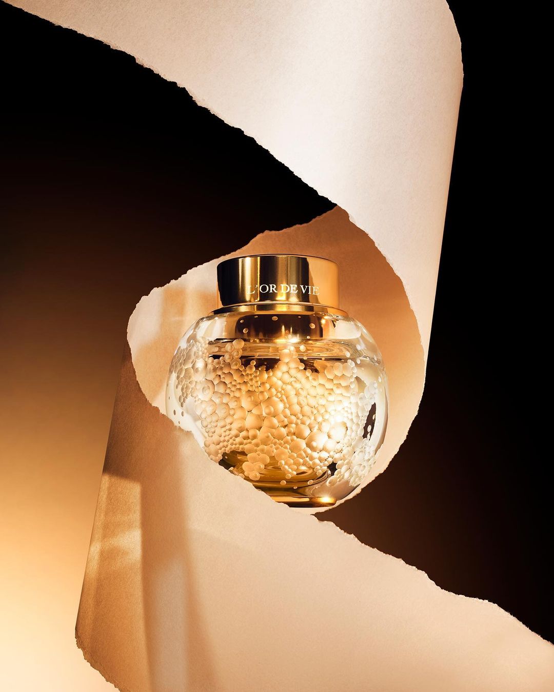 Elegant close-up of L'Or de Vie Dior skincare product in a clear, spherical jar with gold lid, framed by torn paper on a dark backdrop.