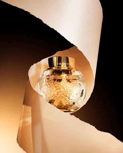 Elegant close-up of L'Or de Vie Dior skincare product in a clear, spherical jar with gold lid, framed by torn paper on a dark backdrop.