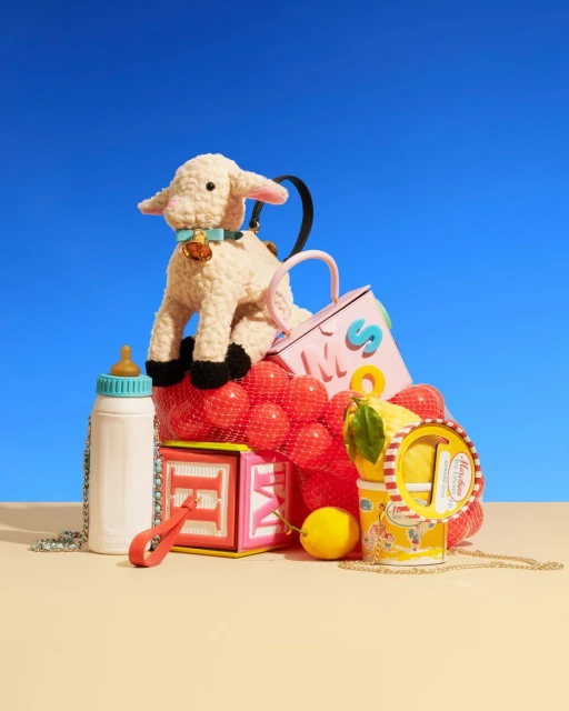 Whimsical handbag arrangement featuring a lamb purse, baby bottle, alphabet blocks, lemons, and red balls against a vibrant blue backdrop.
