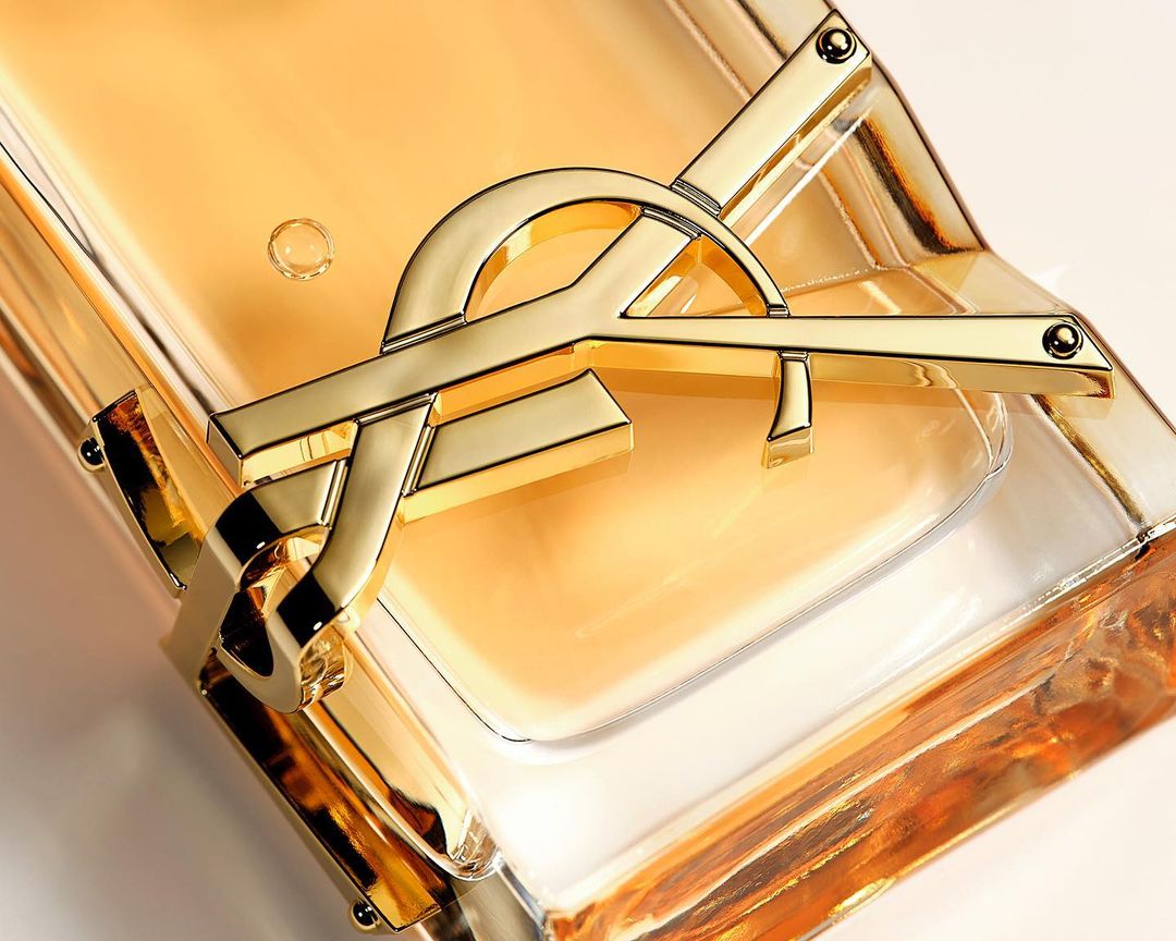Close-up of a gold Yves Saint Laurent logo on a clear glass bottle of amber-colored liquid, set against a light beige background.