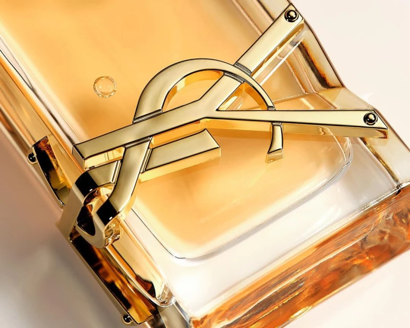 Close-up of a gold Yves Saint Laurent logo on a clear glass bottle of amber-colored liquid, set against a light beige background.