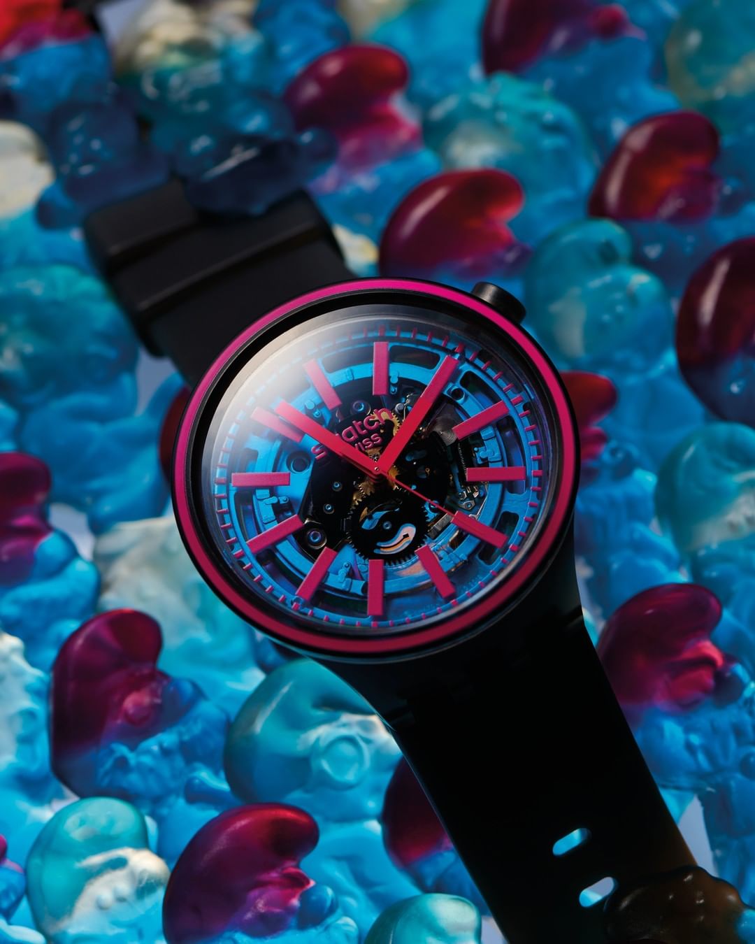 Close-up studio shot of a black wristwatch with a blue and pink face, surrounded by a pile of colorful gummy candies.