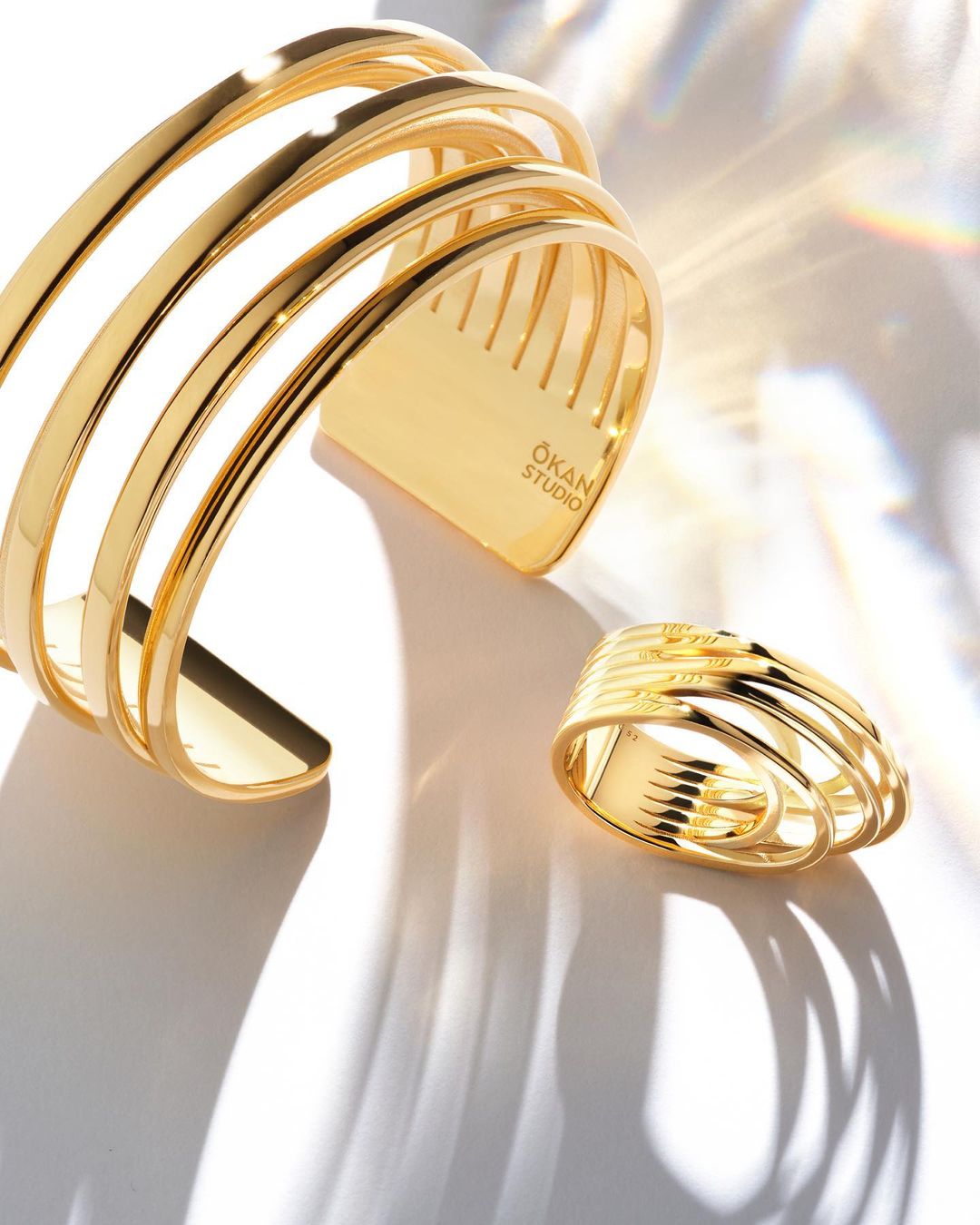 Close-up of a gold ÖKAN STUDIO cuff bracelet and ring on a white surface with light and shadow patterns, showcasing jewelry design.