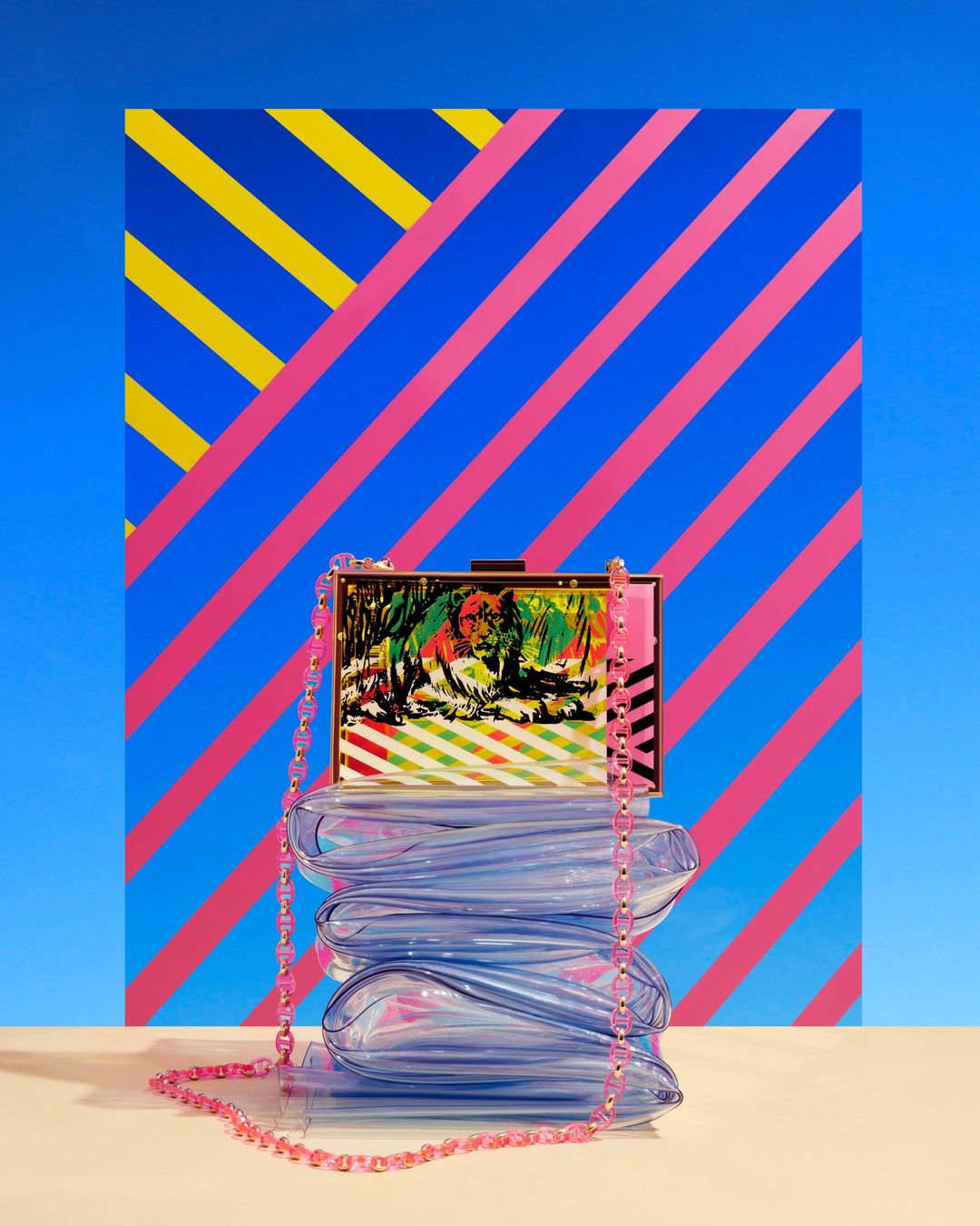 Still life product shot featuring a transparent blue handbag with a pink beaded strap against a vibrant striped backdrop.