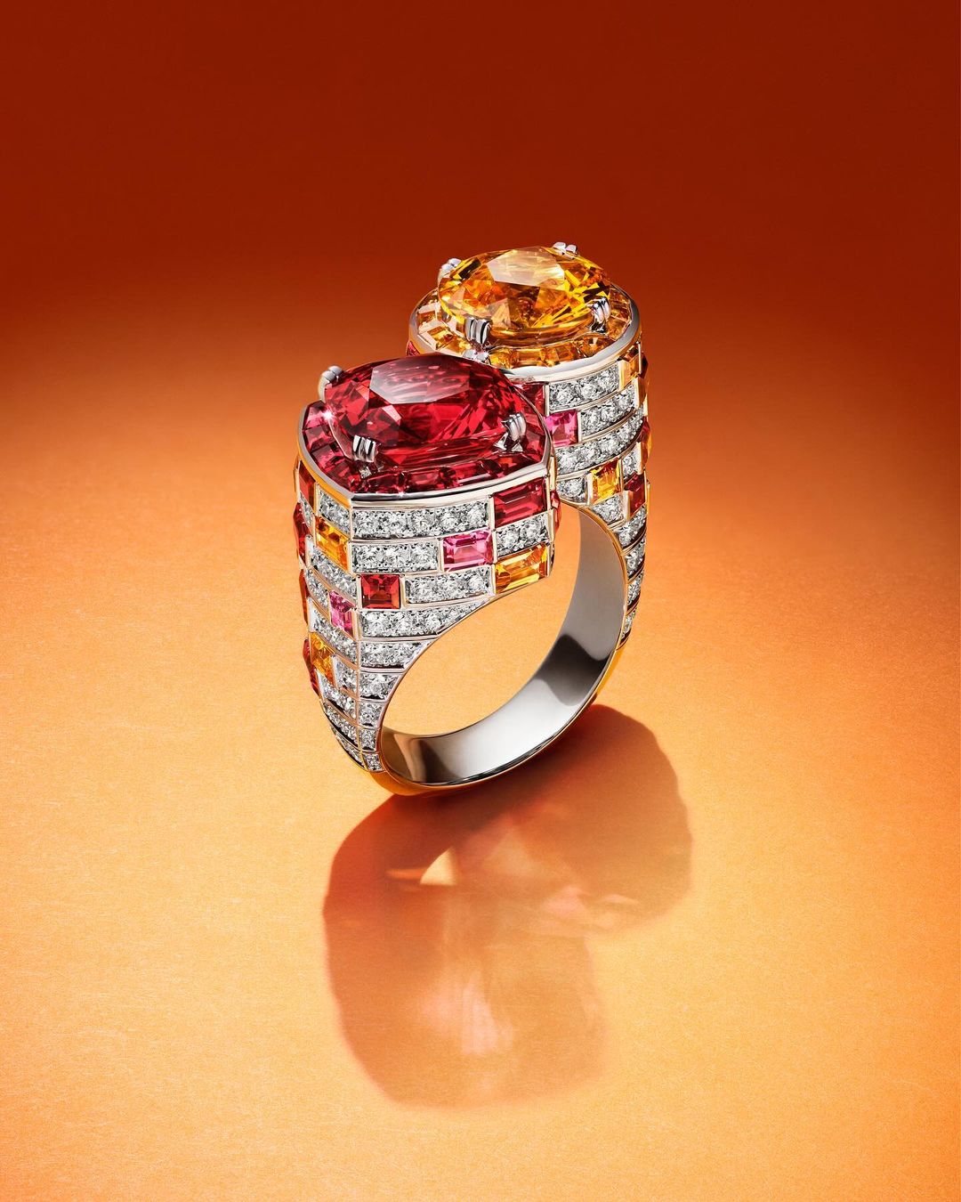 Close-up of a gemstone ring with orange and red stones and diamond accents, set against a warm orange gradient background.