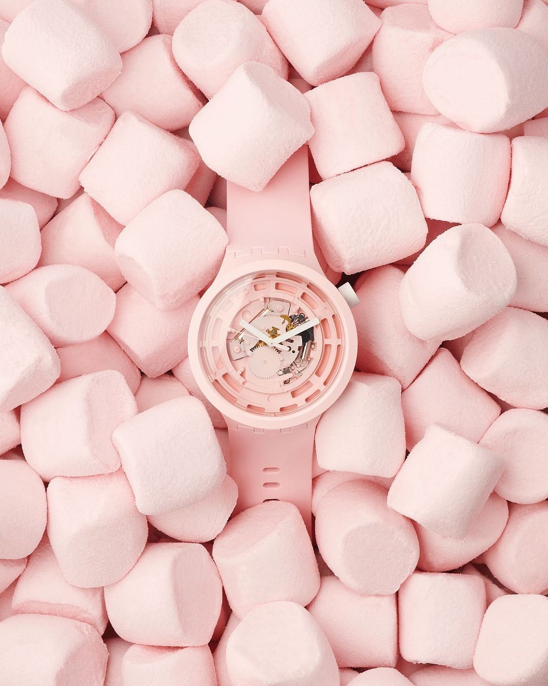 Overhead shot of a light-pink watch lying on a pink marshmallow surface, creating a monochromatic and pastel color scheme.