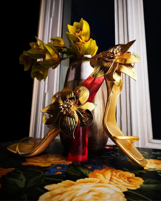 Elegant gold high heels with floral embellishments displayed next to a vase of yellow orchids on a floral-patterned surface.