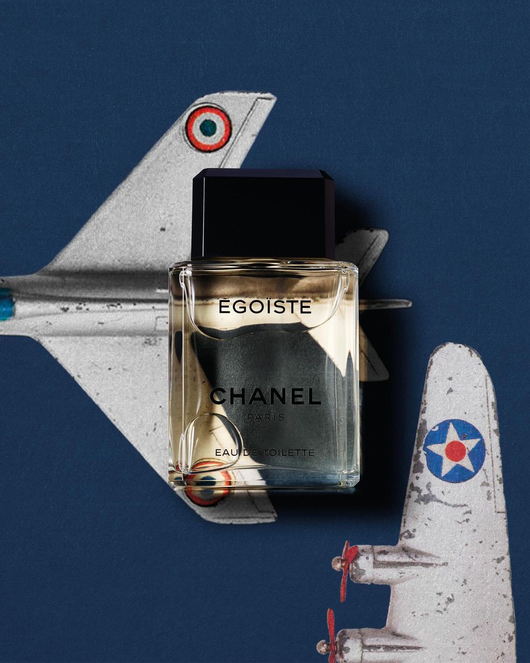 Chanel Egoiste perfume bottle sits in front of two vintage toy airplanes on a dark blue background in a studio shot.
