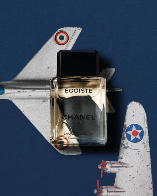 Chanel Egoiste perfume bottle sits in front of two vintage toy airplanes on a dark blue background in a studio shot.