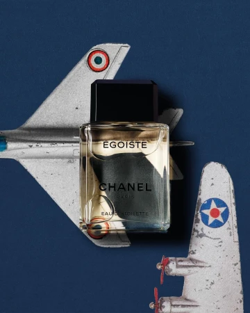 Chanel Egoiste perfume bottle sits in front of two vintage toy airplanes on a dark blue background in a studio shot.