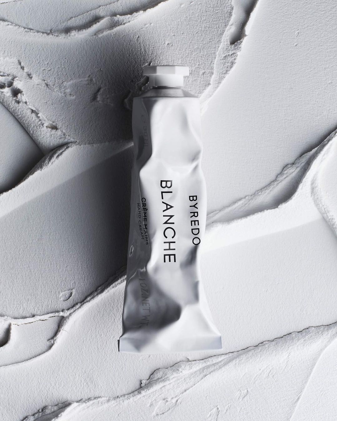 Close-up of Byredo Blanche hand cream tube on textured white background, promoting beauty and personal care products.