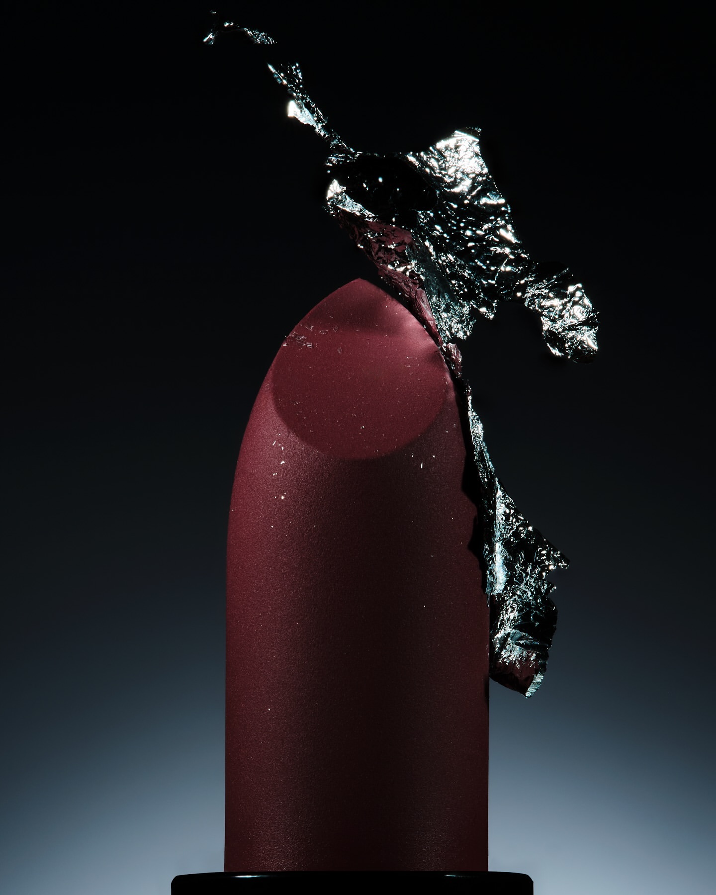 Close-up studio shot of a maroon lipstick tube with silver foil accents against a gradient dark gray background. Beauty product.