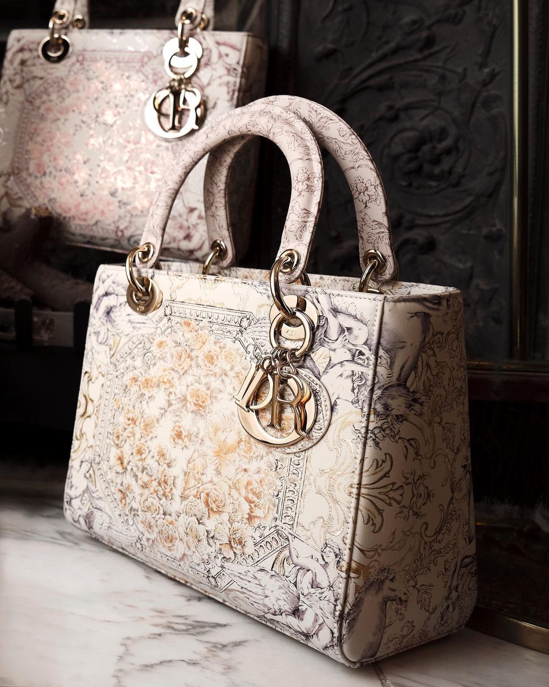 Elegant ivory 'Lady Dior' handbag with ornate print and gold hardware, showcased in a close-up studio shot against a dark textured backdrop.