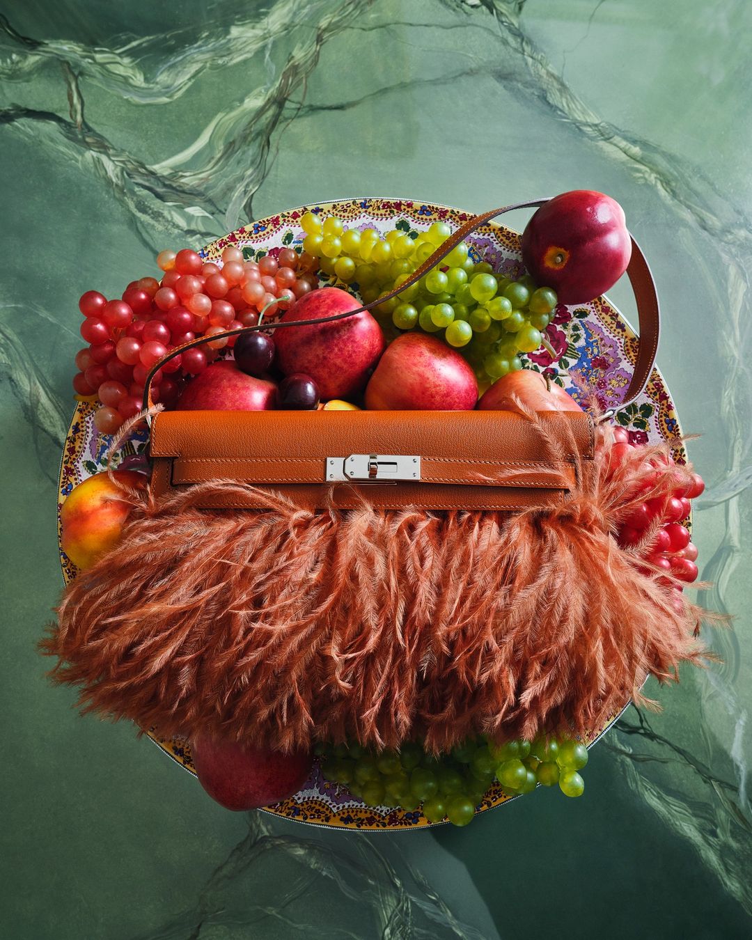 Luxury handbag with fur trim surrounded by fresh fruit displayed on decorative plate against a green marbled background.