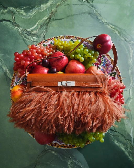 Luxury handbag with fur trim surrounded by fresh fruit displayed on decorative plate against a green marbled background.