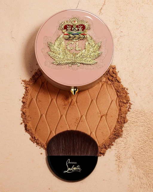 Overhead shot of Christian Louboutin bronzer compact with brush, set on loose bronzer powder on a tan surface.
