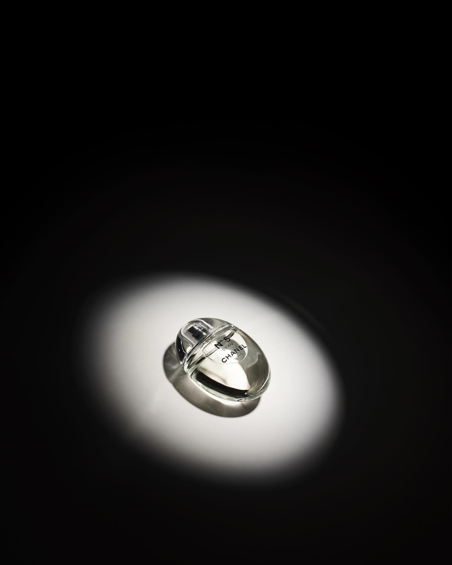 Overhead studio shot of a Chanel perfume bottle in a clear glass bottle, highlighted by a bright spotlight on a black background.