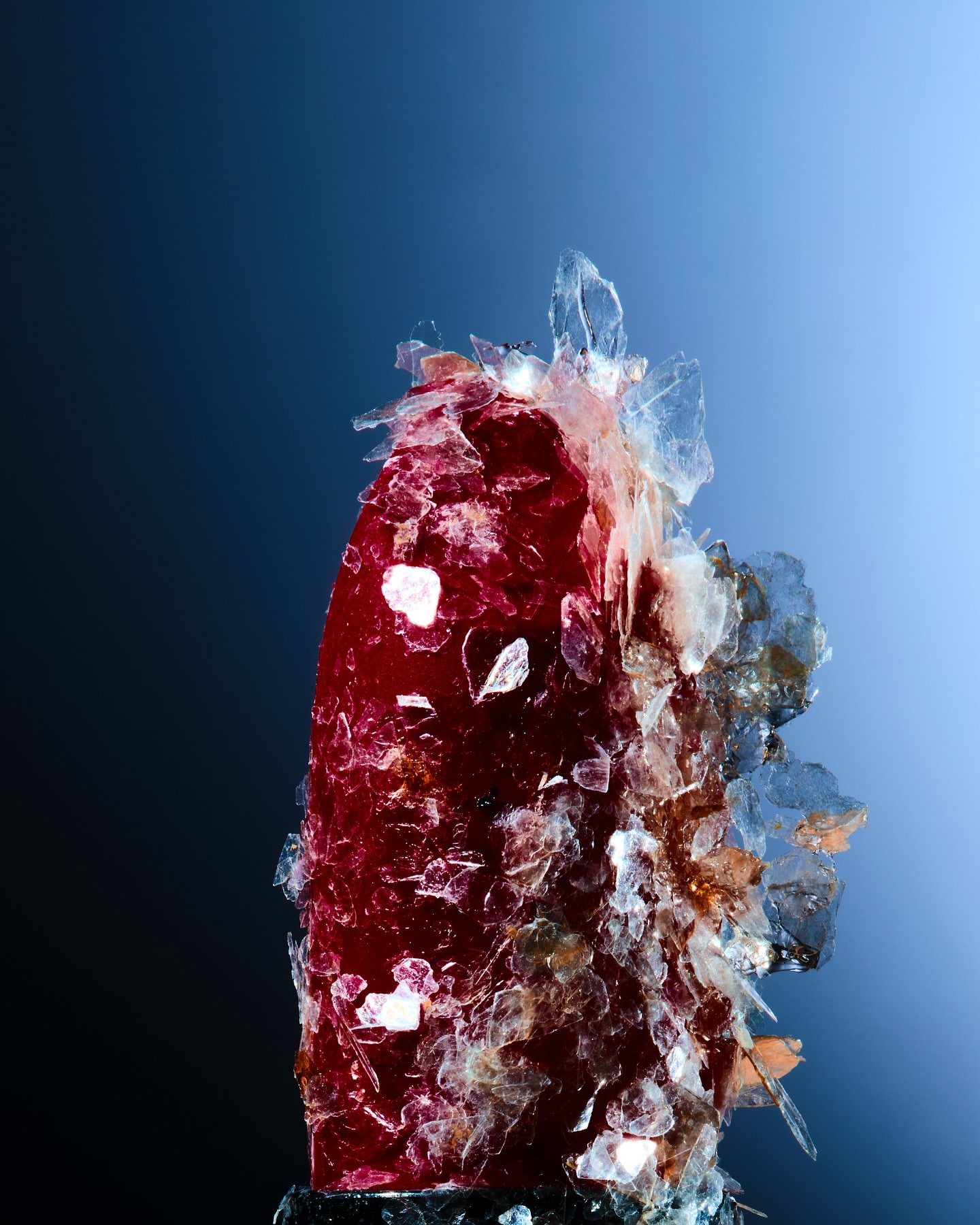 Close-up studio shot of a reddish-brown crystal formation with translucent shards against a dark blue to light blue gradient background.