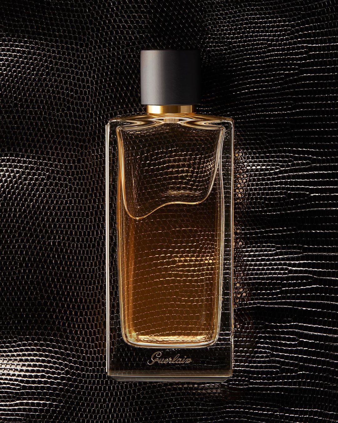 Elegant glass perfume bottle with black cap filled with golden liquid against a shiny black textured surface, close-up product shot.