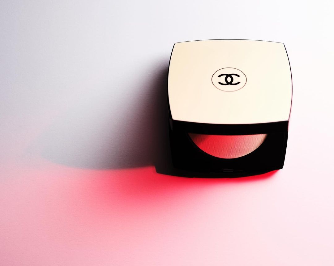 Chanel compact makeup case with pink blush on a white surface illuminated by soft red light in a beauty still-life product shot.