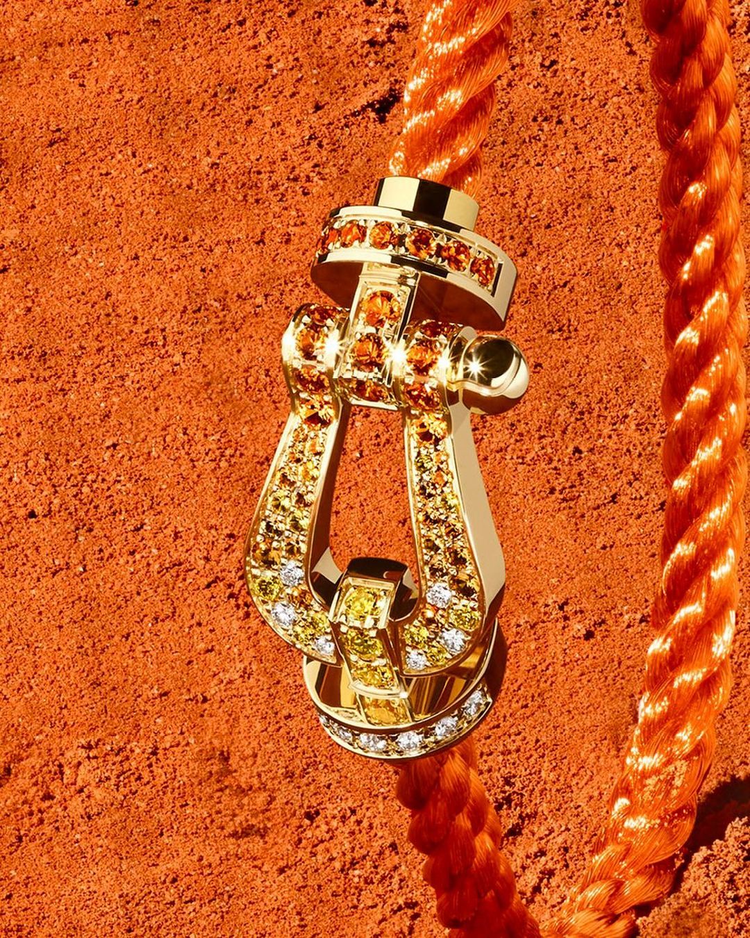 Gold buckle pendant with gem inlays on an orange rope, against an orange textured background, jewelry advertisement close-up.