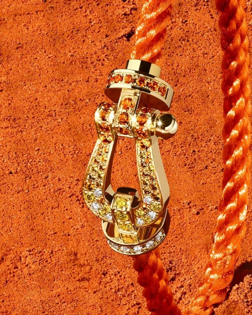 Gold buckle pendant with gem inlays on an orange rope, against an orange textured background, jewelry advertisement close-up.