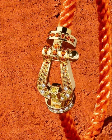 Gold buckle pendant with gem inlays on an orange rope, against an orange textured background, jewelry advertisement close-up.