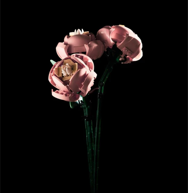 Three pink Lego flower blooms with green stems on a black background. The flowers are lit from above, creating a soft, diffused light.