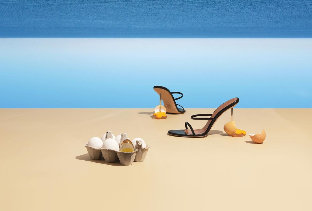 High heels and egg yolks create a surreal fashion still life on a seamless tan surface with ocean backdrop for beauty or advertising.