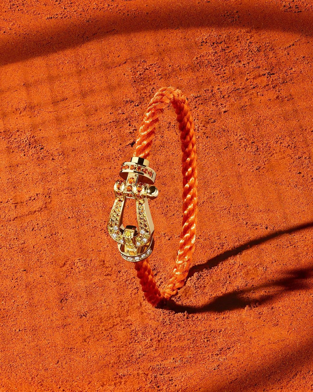 Braided orange bracelet with gold clasp and gems showcased on a textured orange surface with soft shadows, jewelry still life.