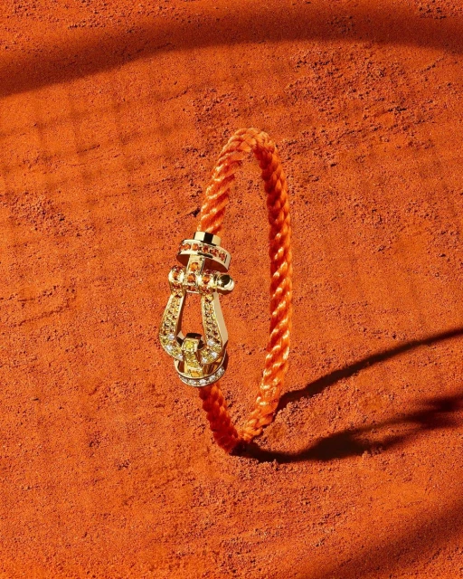 Braided orange bracelet with gold clasp and gems showcased on a textured orange surface with soft shadows, jewelry still life.