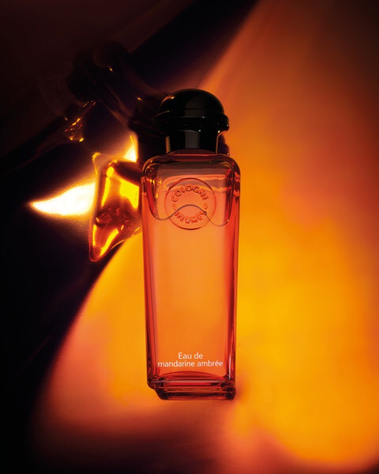 Cologne bottle of Eau de mandarine ambrée by Hermès, studio shot on orange gradient backdrop with metal design reflecting light.