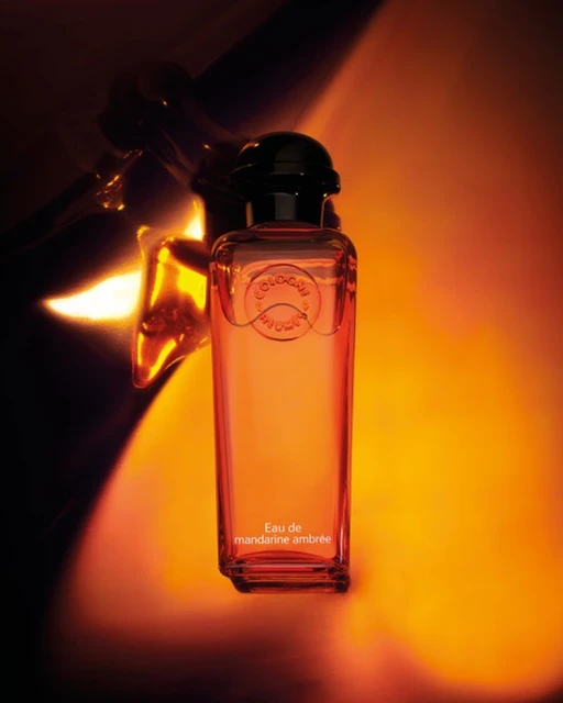 Cologne bottle of Eau de mandarine ambrée by Hermès, studio shot on orange gradient backdrop with metal design reflecting light.
