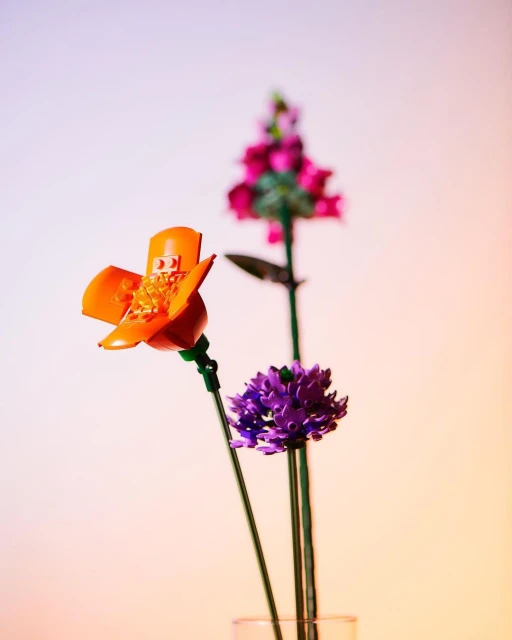 Three colorful Lego flowers, including an orange California poppy and purple allium, are arranged in a clear vase with a soft gradient background.
