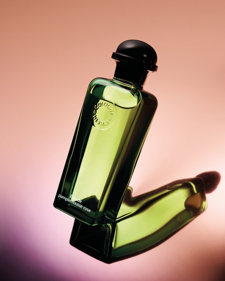Studio shot of a green glass Hermès cologne bottle with a black cap, angled on a reflective surface with a peach to lavender gradient background.