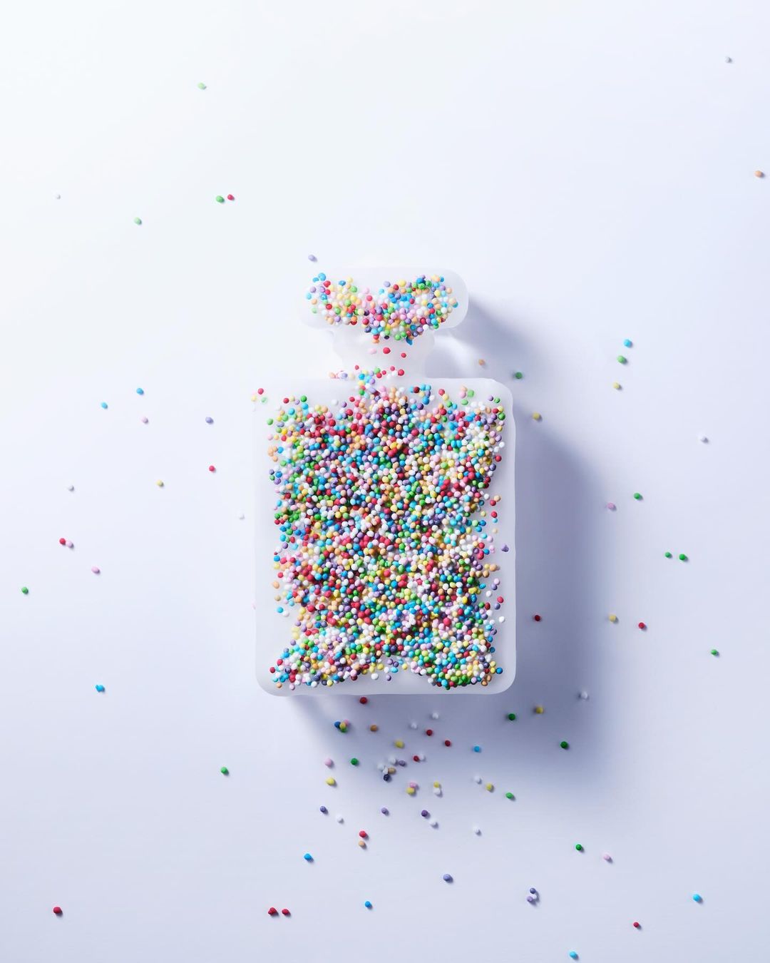 Overhead shot of a clear perfume bottle silhouette filled with multicolored sprinkles on a bright white background, minimalist concept.
