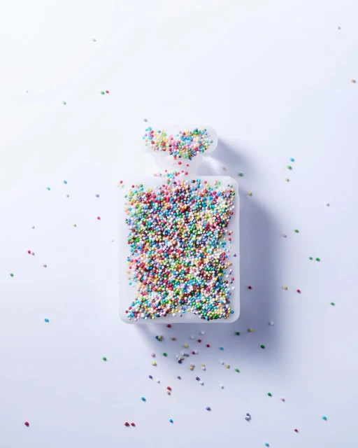 Overhead shot of a clear perfume bottle silhouette filled with multicolored sprinkles on a bright white background, minimalist concept.