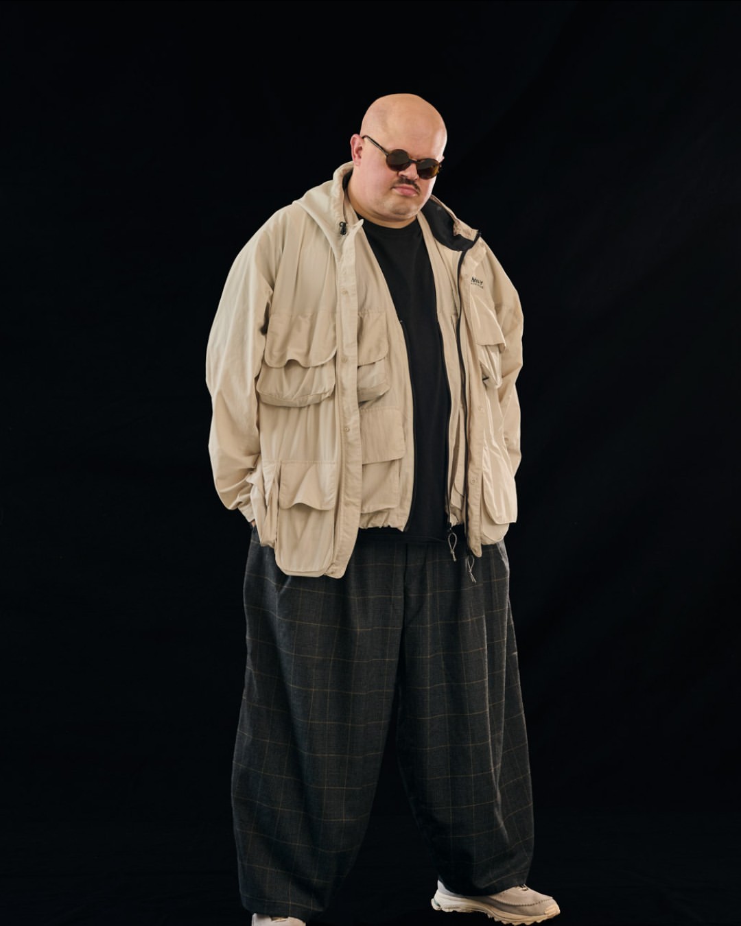 Full shot of a bald man with sunglasses, wearing a black t-shirt, beige jacket, and checkered pants, standing in front of a black backdrop.